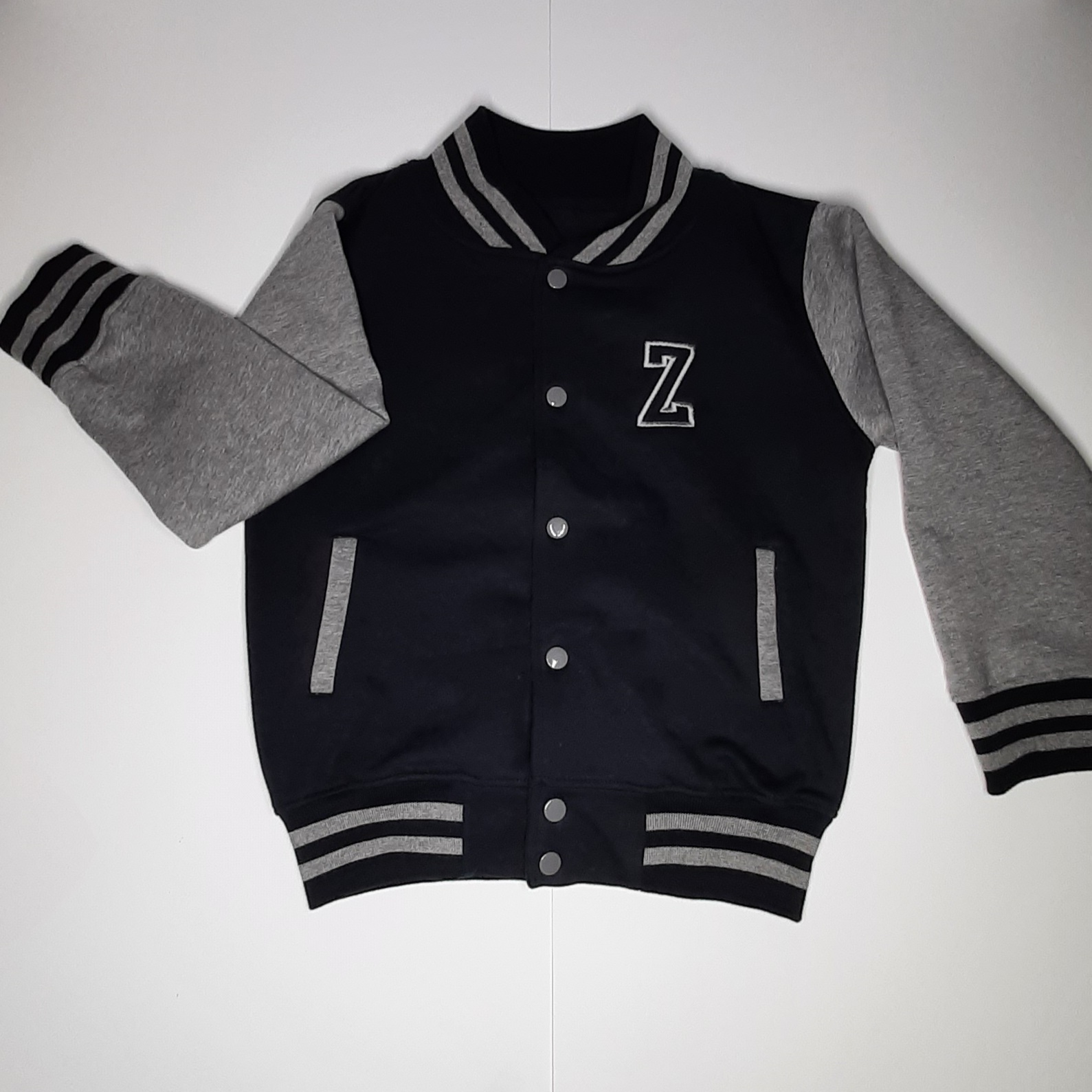 Children's varsity jacket on model in black and grey with embroidered initial laid out