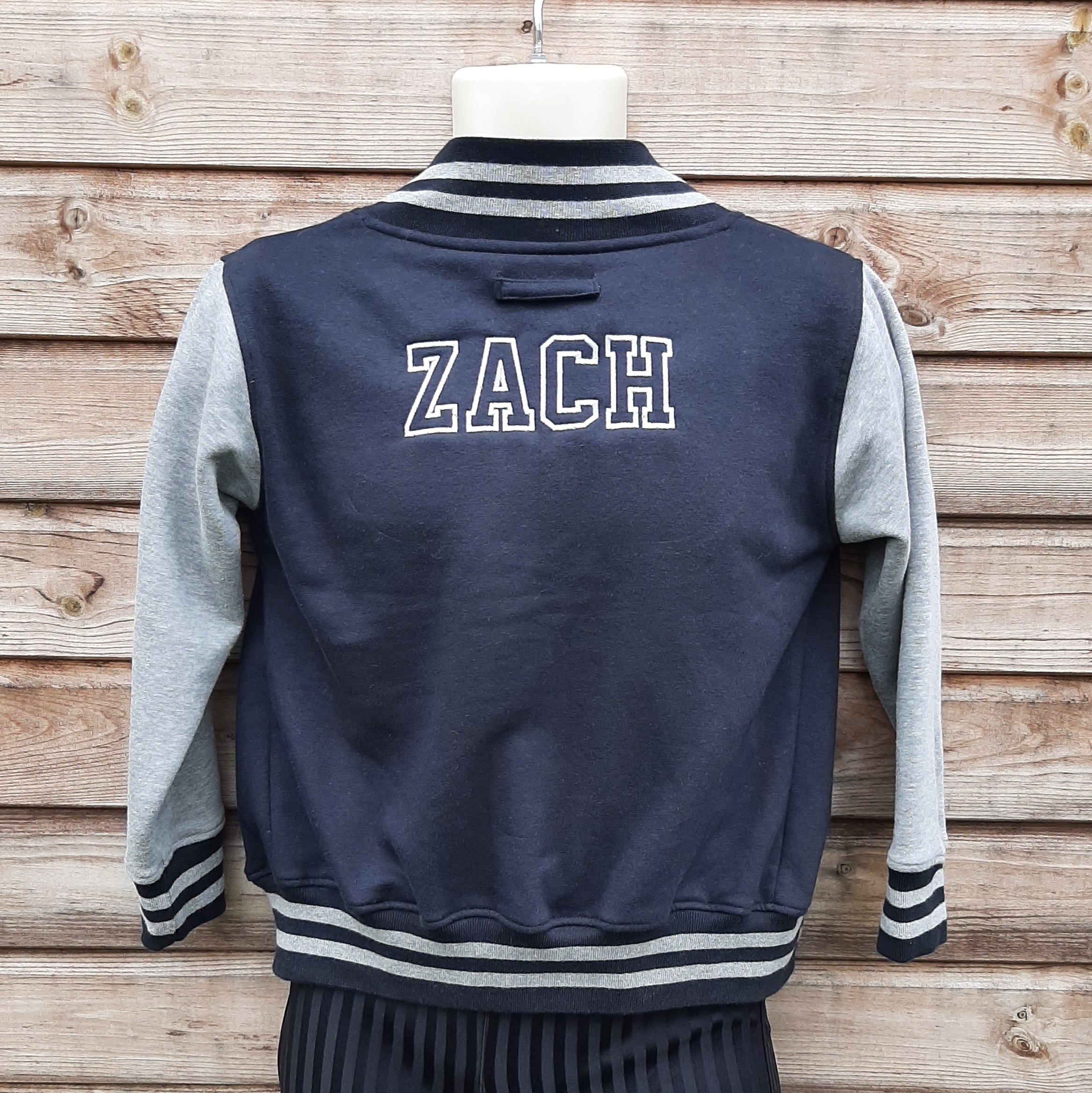 Children's varsity jacket on model in black and grey with embroidered name on the back hung up on model